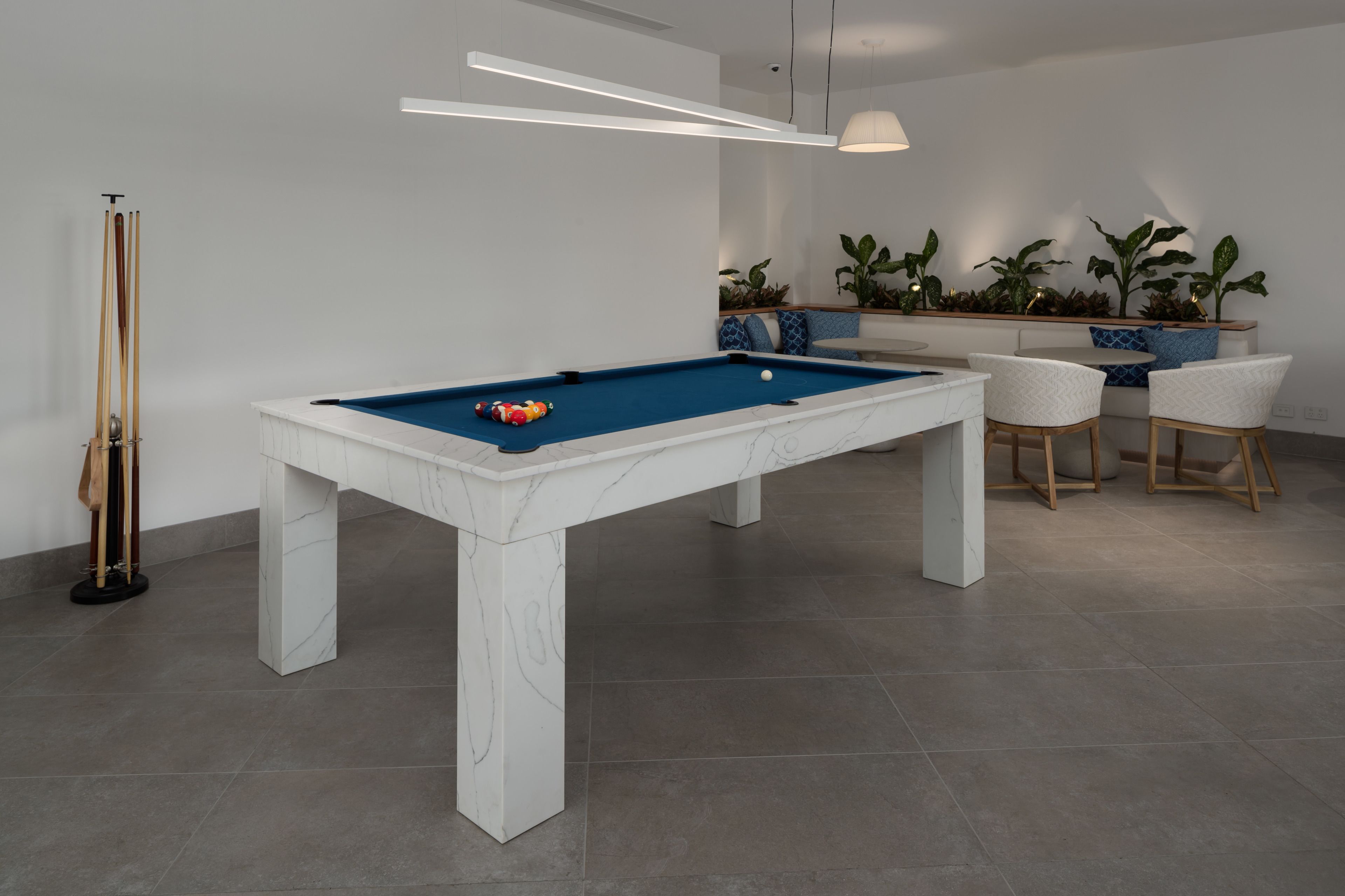 Una mesa de billar con superficie de fieltro azul, rodeada por una base de mármol blanco. Hay dos sillas blancas con patas de madera y una mesa pequeña con cojín azul. Se ve una planta en maceta al fondo.