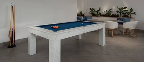 Una mesa de billar con superficie de fieltro azul, rodeada por una base de mármol blanco. Hay dos sillas blancas con patas de madera y una mesa pequeña con cojín azul. Se ve una planta en maceta al fondo.