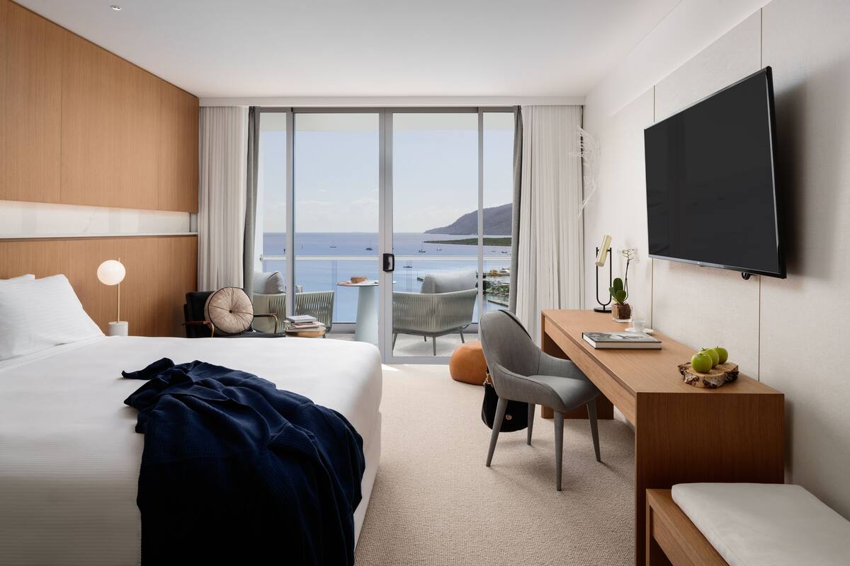 A hotel room with a large bed, a desk, a chair, a television, and a view of the sea.