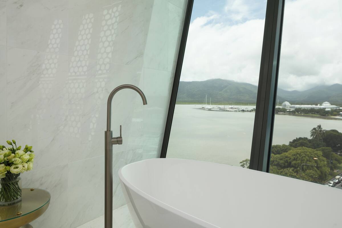 A modern bathroom with a freestanding bathtub, a glass vase with flowers, and a view of a body of water and mountains through large windows.
