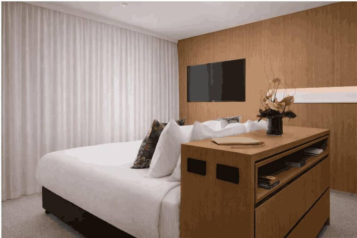 A modern bedroom with a large bed, a wooden dresser, and a mounted TV.
