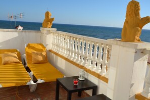 Een balkon met een witte balustrade, twee leeuwenbeelden, een gele bank, een kleine tafel met een kaars, en uitzicht op zee.