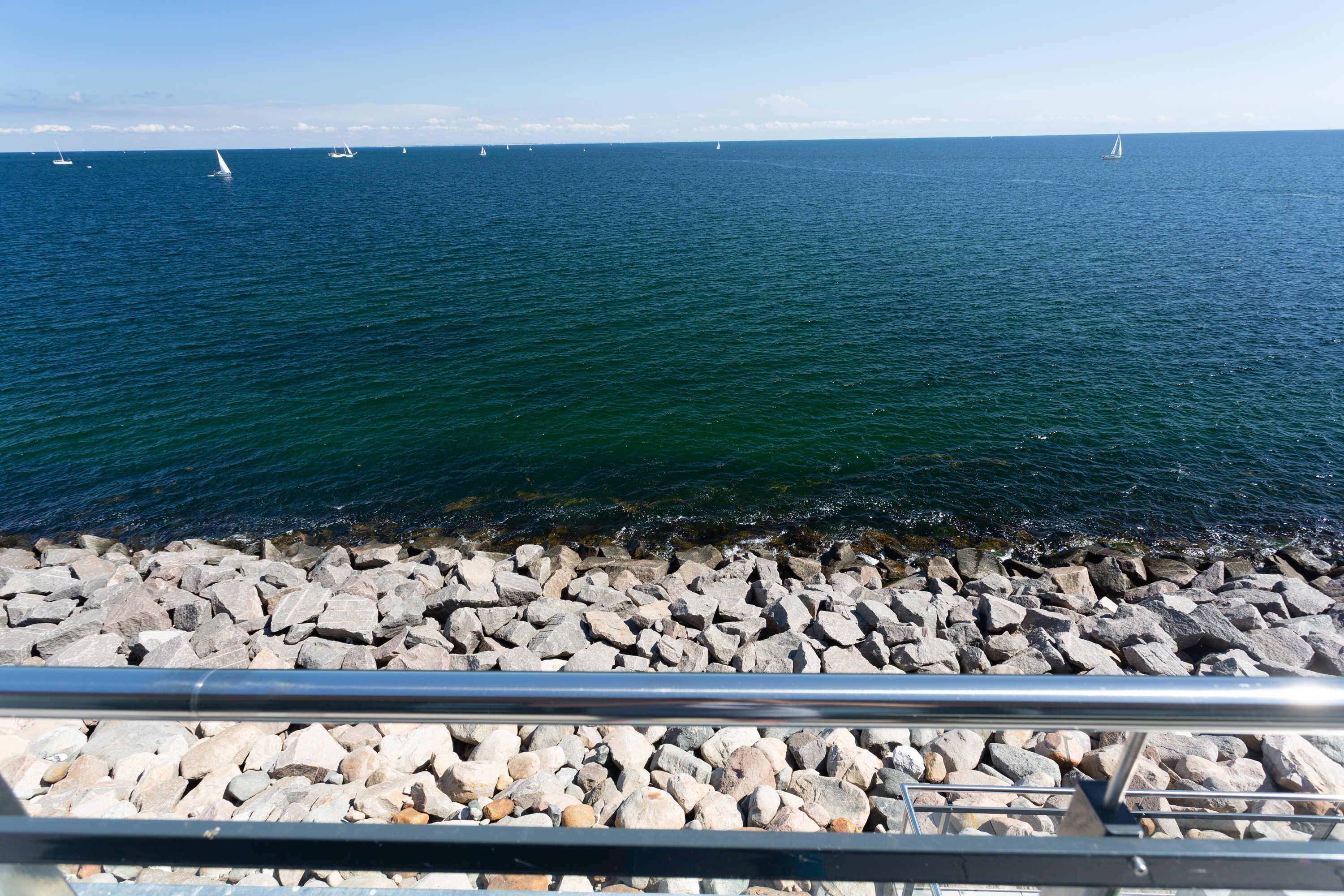 A rocky shoreline with a railing in the foreground and sailboats on the water in the background.