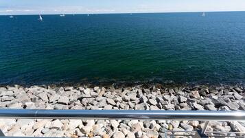 A rocky shoreline with a railing in the foreground and sailboats on the water in the background.