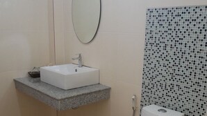 Une salle de bain avec des toilettes blanches, un lavabo rectangulaire surmonté d’un miroir et une zone de douche carrelée.