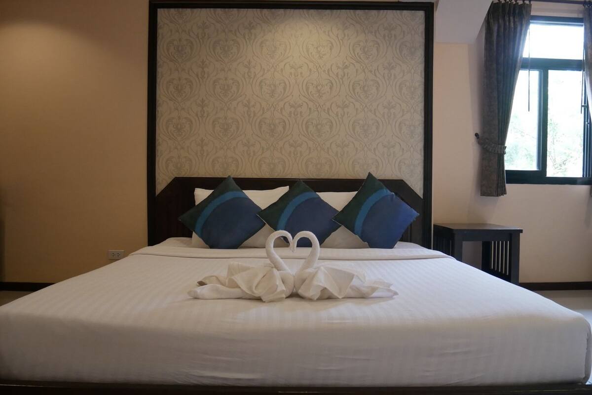 Una cama bien hecha con un arreglo de almohadas en forma de cisne, un cabecero estampado y una ventana con cortinas.