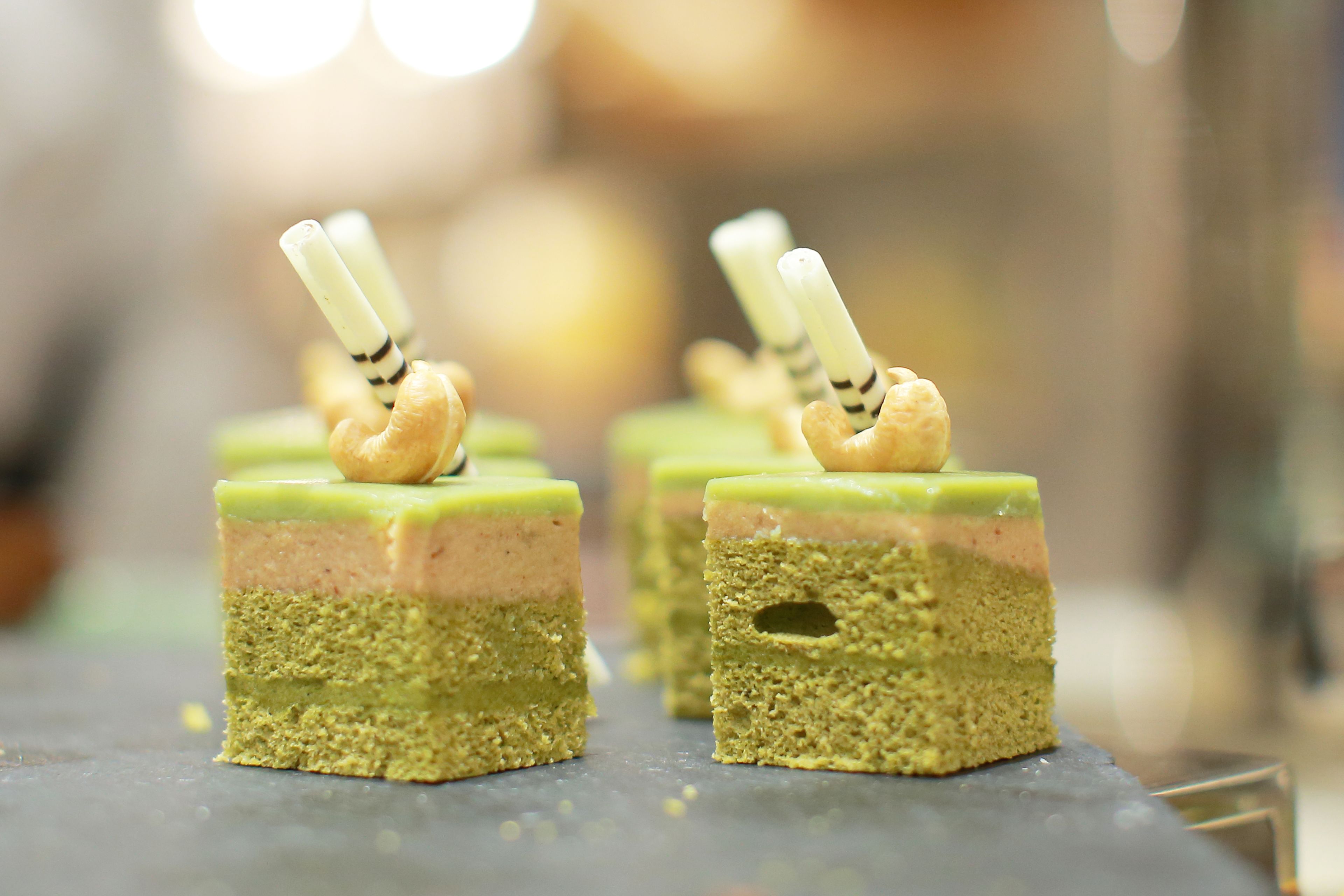 A close-up of square-shaped desserts with a green top layer, a middle layer, and a bottom layer, each topped with a white chocolate stick and a nut.