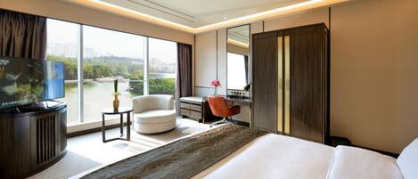 A modern hotel room with a large bed, a desk, a chair, a television, and a view of a cityscape through large windows.