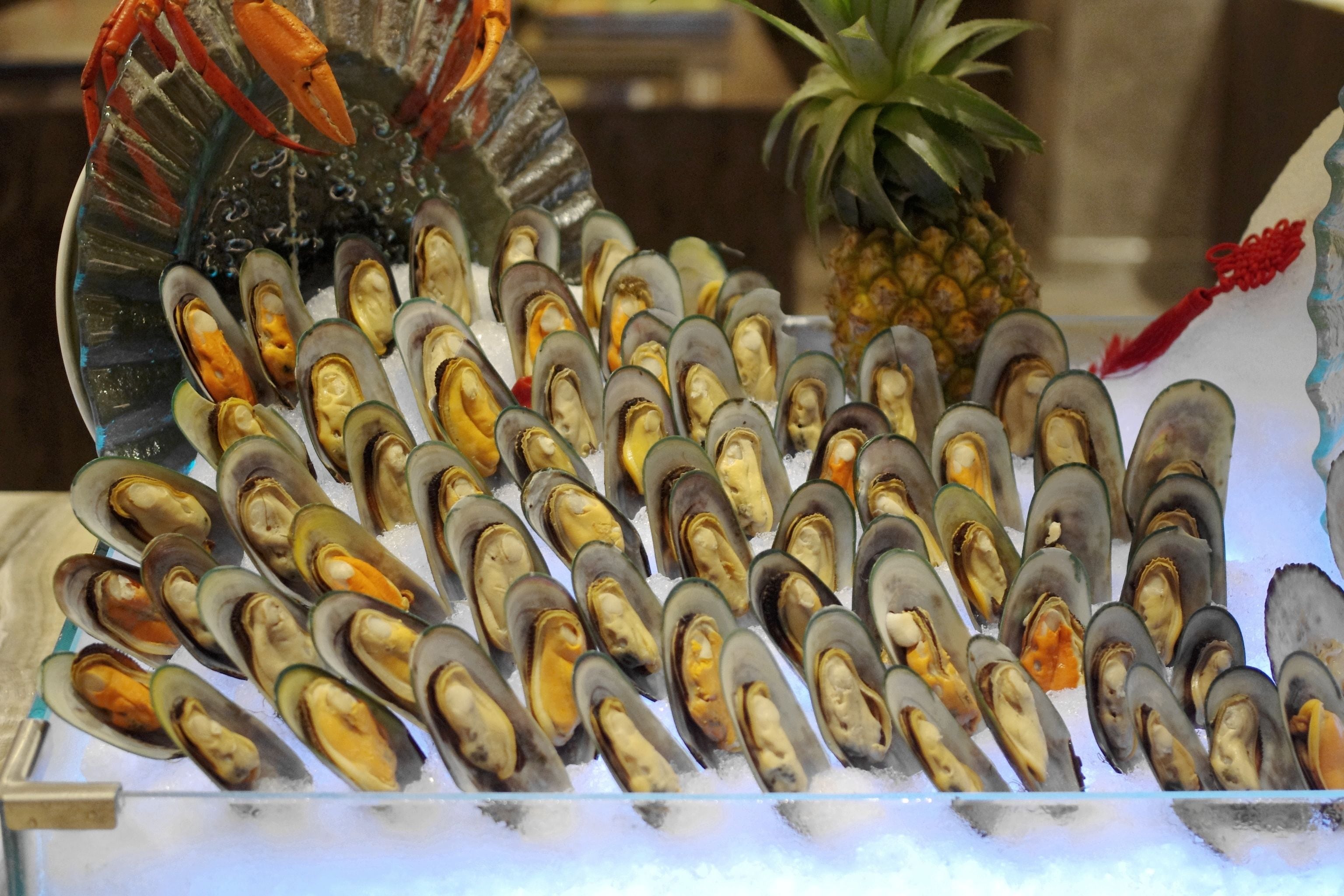 A display of mussels on ice with a lobster and pineapple in the background.