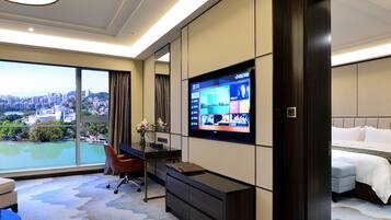 Habitación de hotel con un televisor grande montado en la pared, un escritorio con una silla, una cama y vistas a un paisaje urbano a través de amplias ventanas.