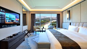 Habitación de hotel moderna con una televisión grande, un sofá, una cama y vistas a un paisaje urbano a través de ventanas amplias.
