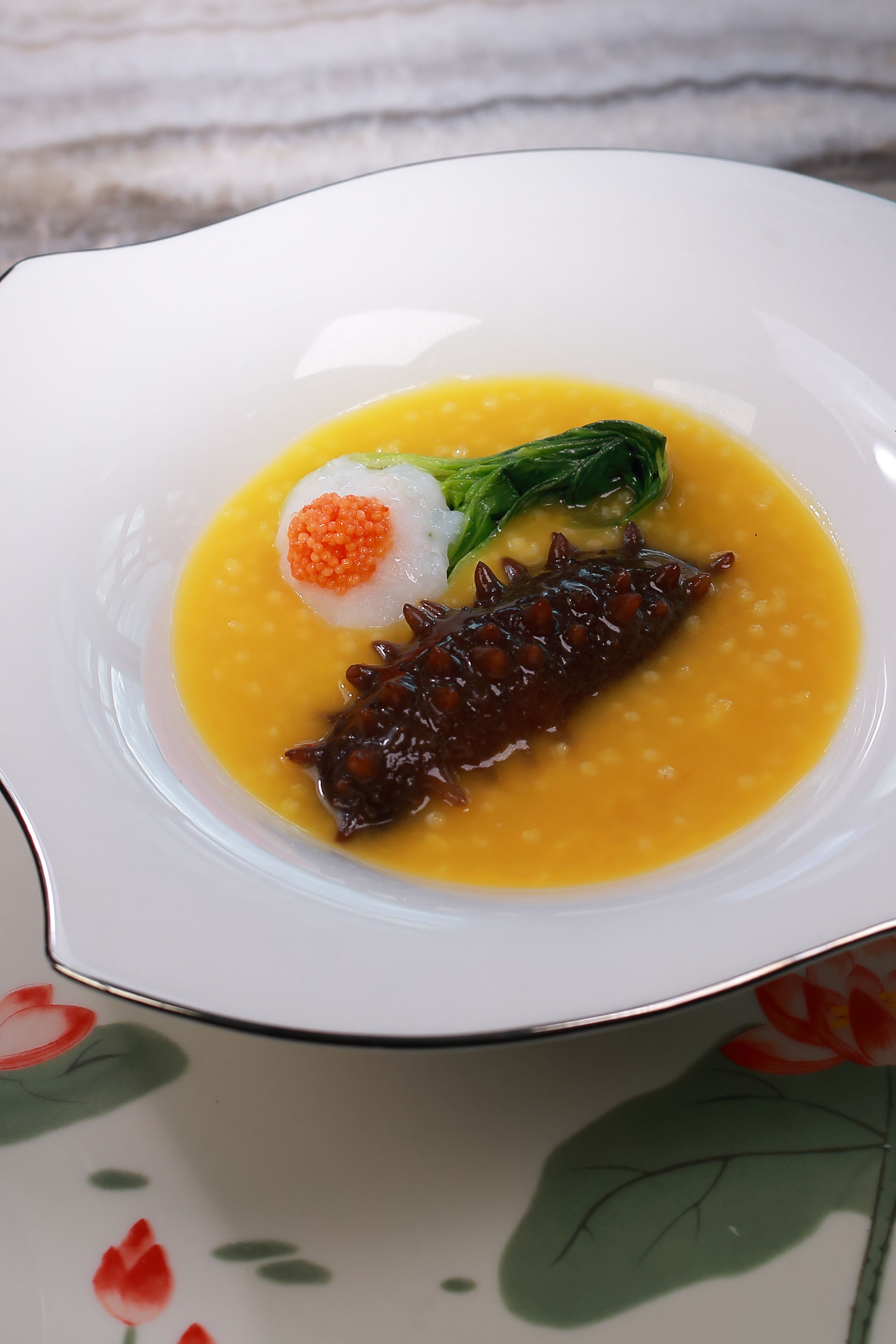 Une assiette blanche ornée d’un motif floral, contenant un plat de soupe jaune, une boulette blanche garnie de caviar orange et un champignon foncé.