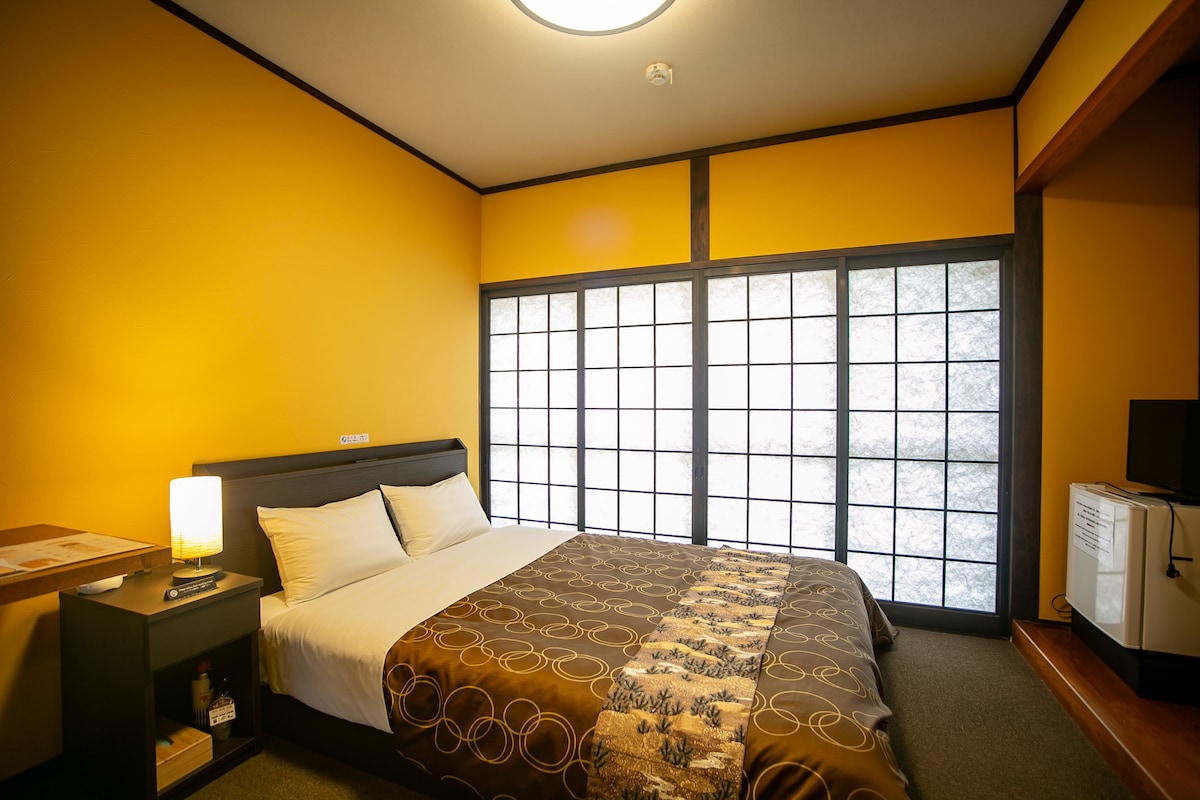 Una habitación de hotel con un ventanal grande, una cama con colcha estampada, una mesita de noche con lámpara y un televisor pequeño.