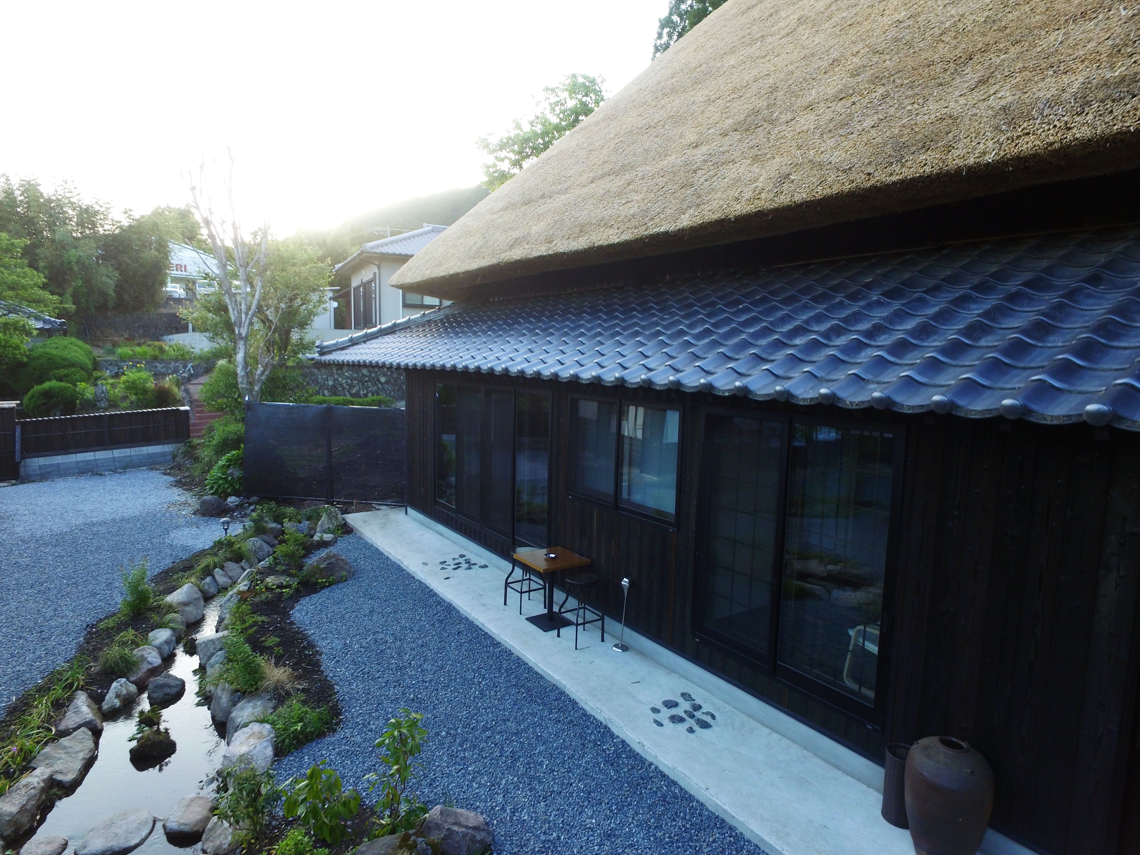 Une maison traditionnelle de style japonais, avec un toit de chaume, un chemin de gravier et un petit espace extérieur aménagé avec une table et des chaises.