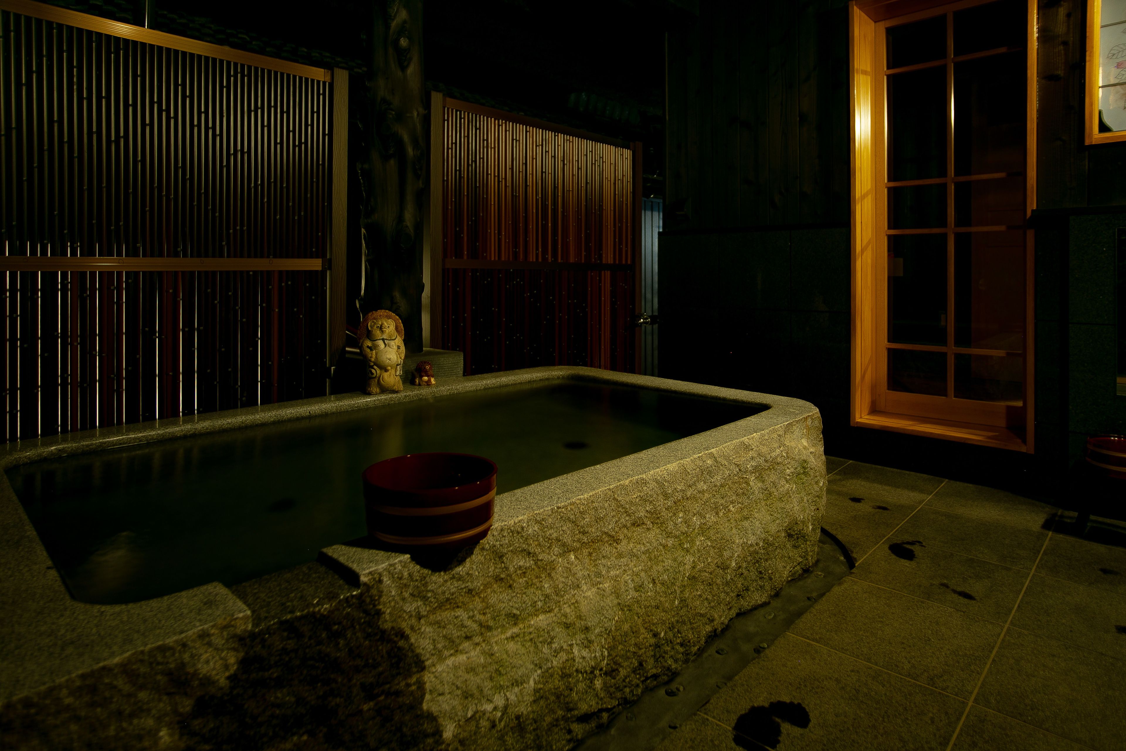 A traditional Japanese onsen with a stone bath, wooden sliding doors, and a small Buddha statue.