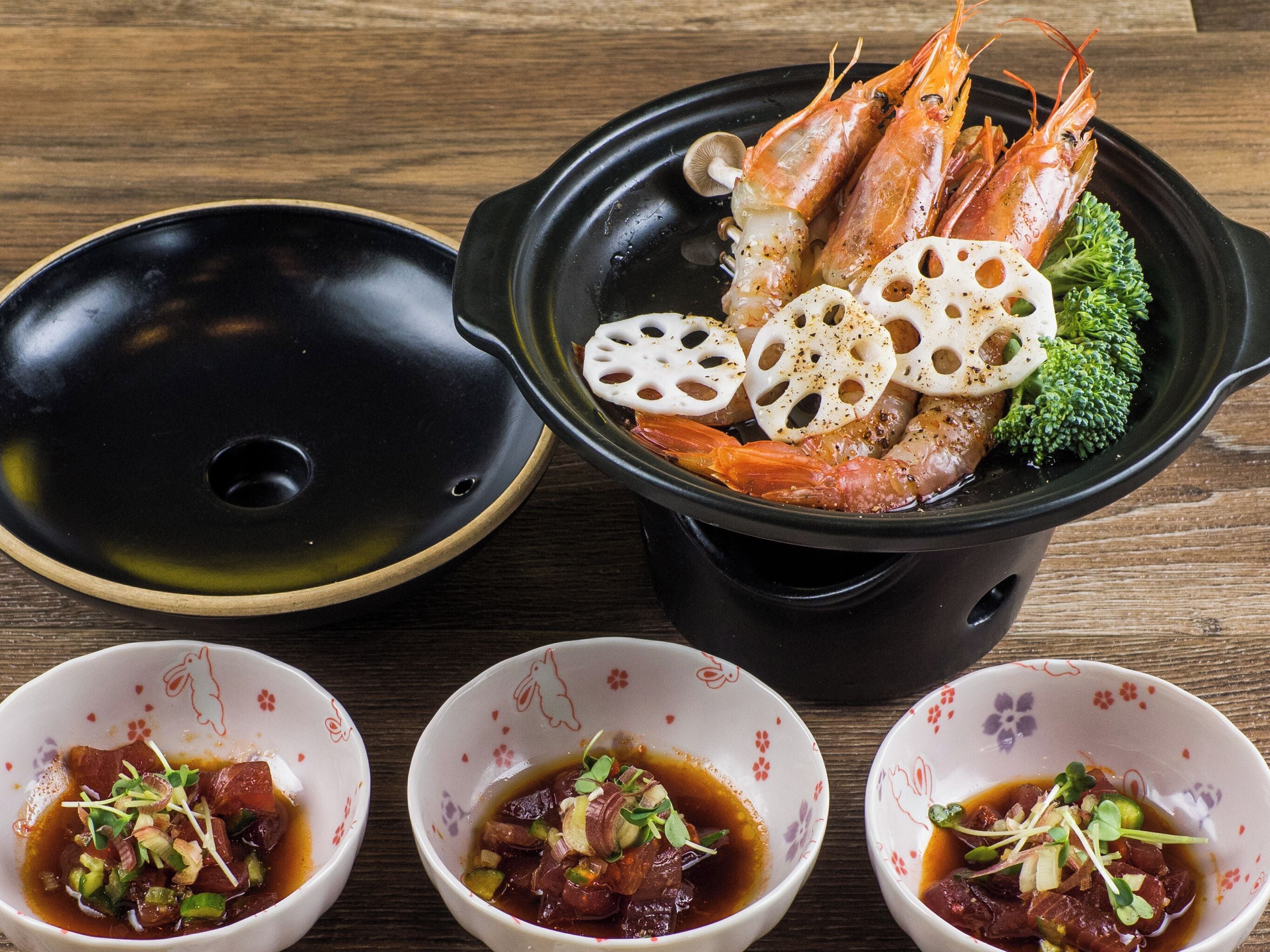 Una mesa con un recipiente para fondue, tres tazones de sopa y un plato con camarones y verduras cocidas.