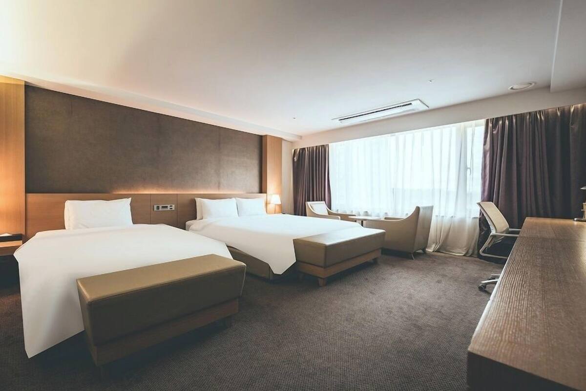 Une chambre d’hôtel avec deux lits, un bureau, une chaise et une grande fenêtre avec des rideaux.