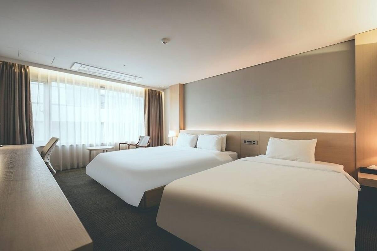 Une chambre d’hôtel avec deux lits, un bureau, une chaise et une grande fenêtre avec des rideaux.