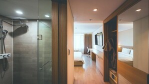 Ein modernes Hotelzimmer mit einer gläsernen Duschkabine, einem Holztischchen mit Spiegel und einem Bett mit weißer Bettwäsche sowie einem begehbaren Kleiderschrank.
