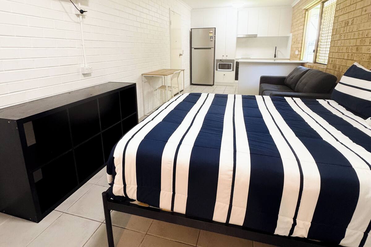 A compact hotel room with a bed, a TV mounted on the wall, a ceiling fan, a kitchenette, and a window with a view.