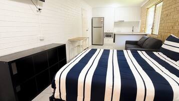 A compact hotel room with a bed, a TV mounted on the wall, a ceiling fan, a kitchenette, and a window with a view.