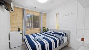 A bedroom with a bed, a small white refrigerator, a window with blinds, a potted plant, and a white wall with hooks. - Motel Sunshine Coast (Caloundra)
