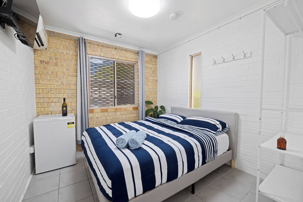 A bedroom with a bed, a small white refrigerator, a window with blinds, a potted plant, and a white wall with hooks.