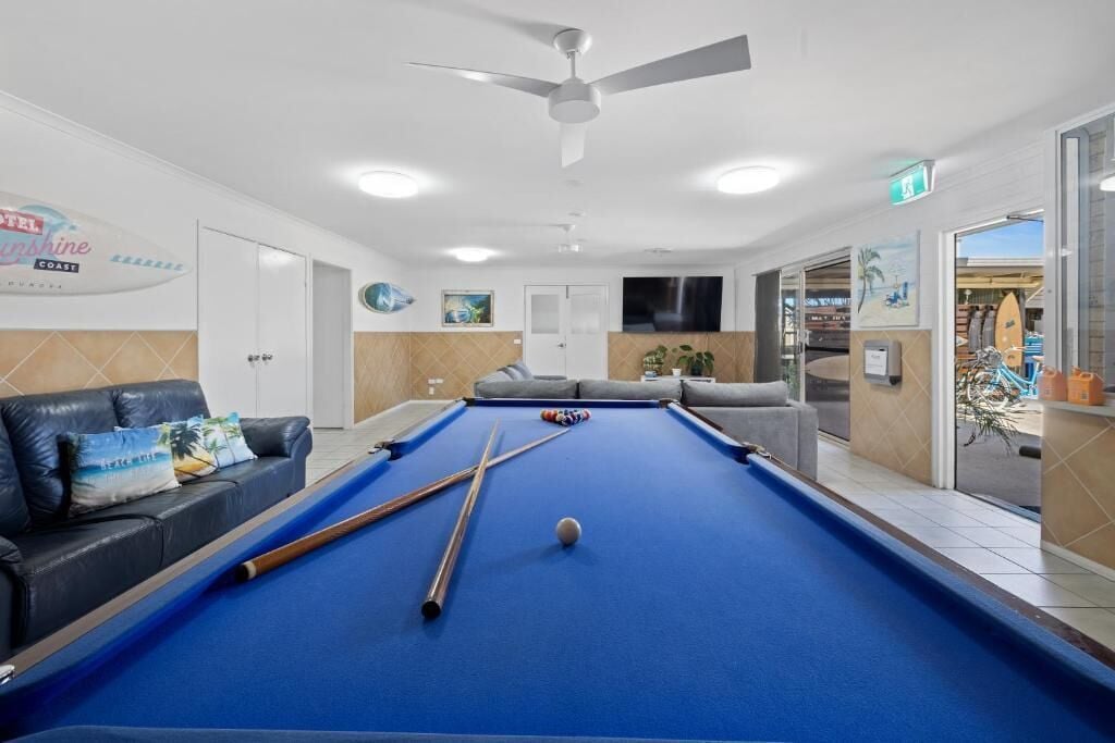 A pool table with pool cues and a ball, a sofa with cushions, a ceiling fan, and a sign for 'Hotel Sunshine Coast'.