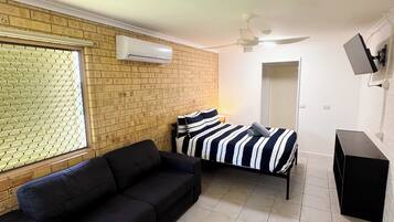 A room with a black sofa, a bed with striped bedding, a tiled floor, a brick wall, and a window with blinds.