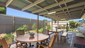 An outdoor patio area with a round wooden table and chairs, a covered seating area, and a garden with various plants.