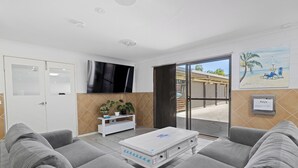 A living room with grey sofas, a white coffee table, a TV, a sliding glass door to the outside, and a painting of a beach scene. - Motel Sunshine Coast (Caloundra)
