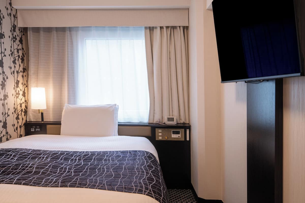 A hotel room with a bed, a bedside table with a lamp, a digital control panel, and a television mounted on the wall.