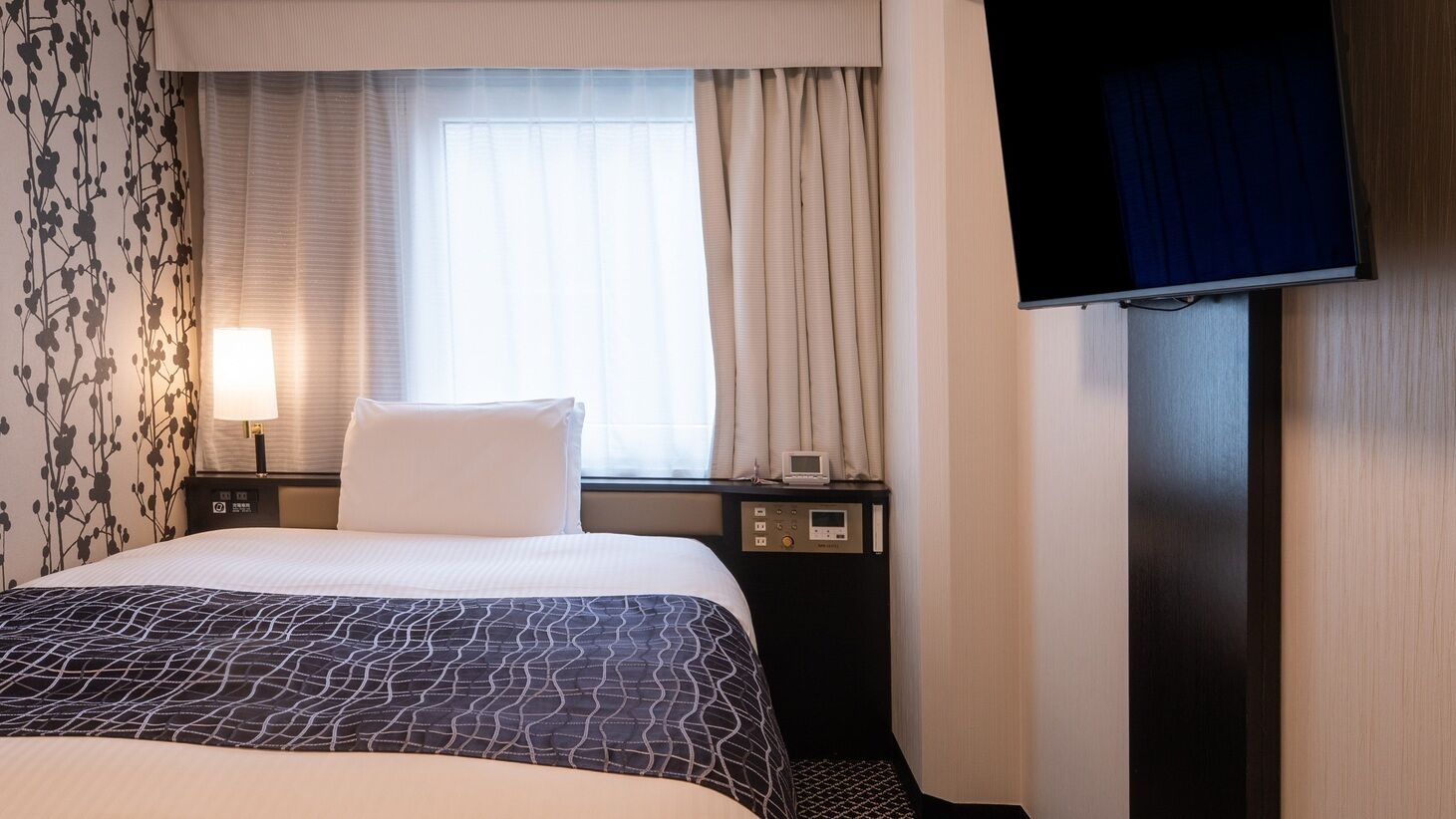 A hotel room with a bed, a TV, a bedside table with a lamp, a window with curtains, and a wall with a floral pattern.
