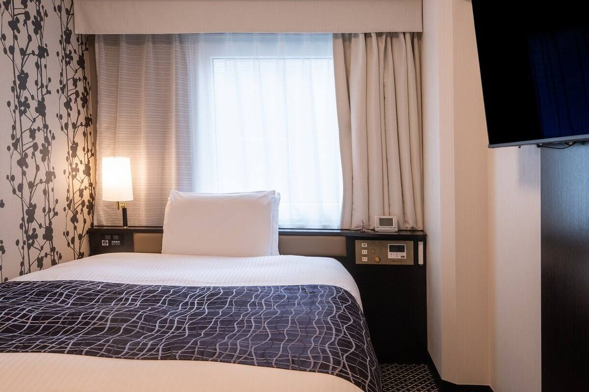 A hotel room with a bed, a TV, a bedside table with a lamp, a window with curtains, and a wall with a floral pattern.
