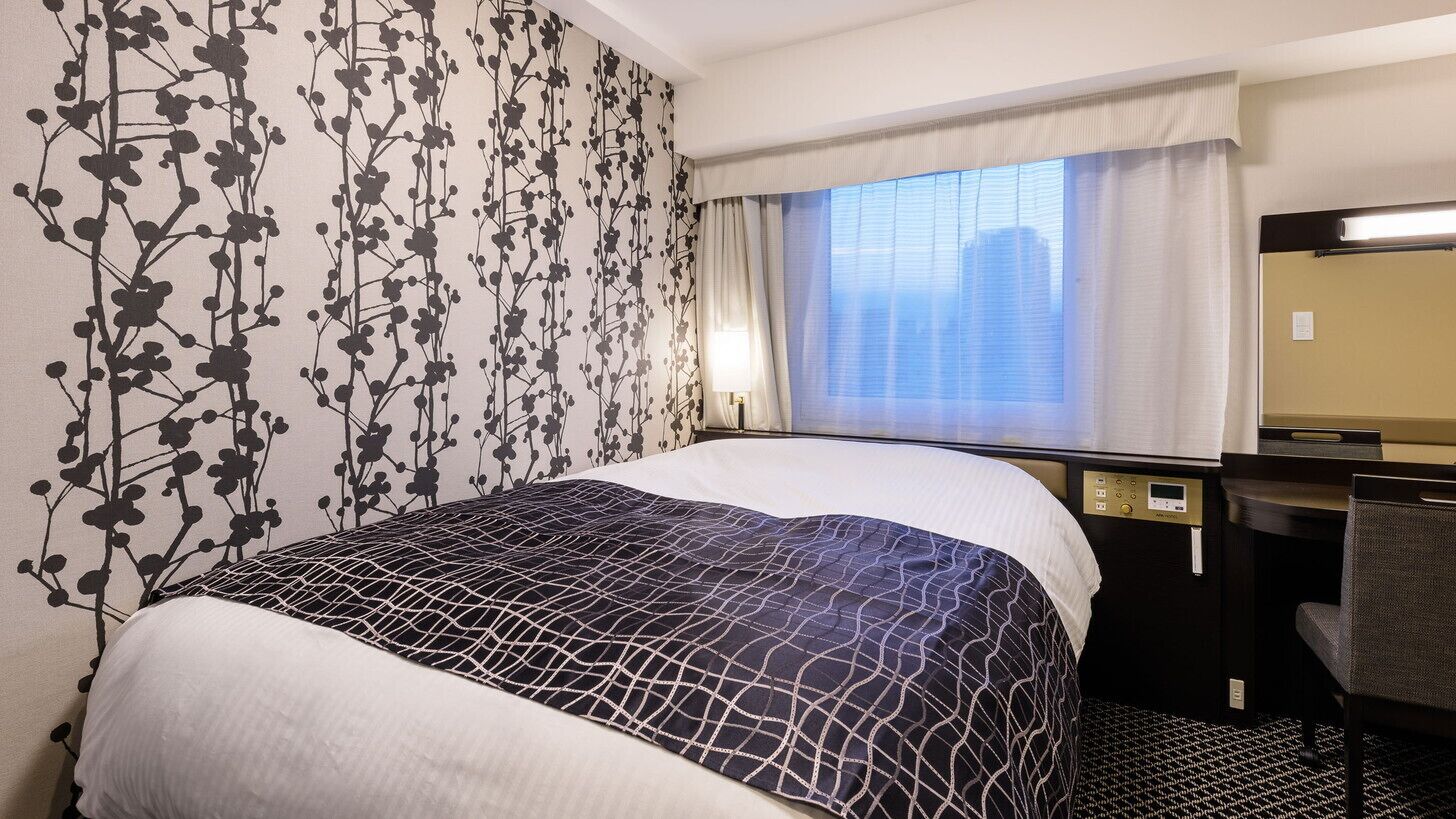 A hotel room with a bed, a desk, a chair, a window with curtains, and a wall with a floral pattern.
