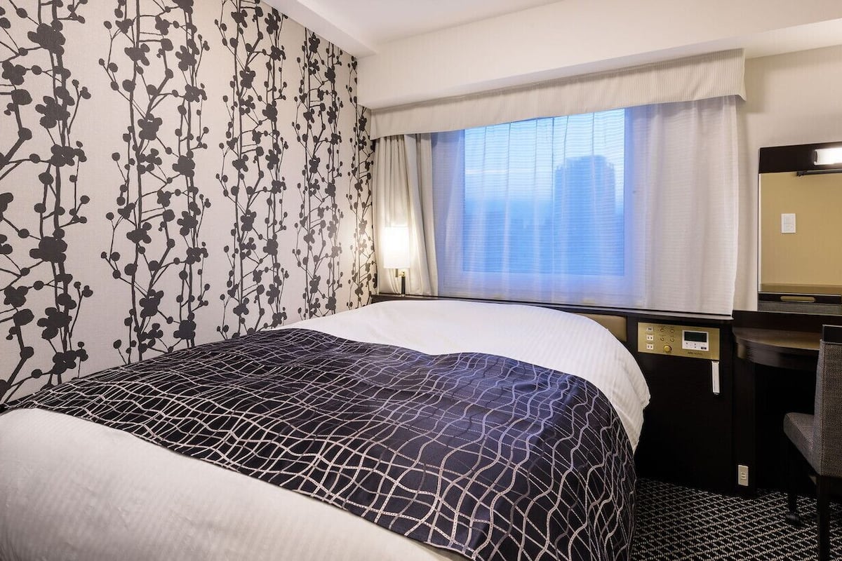A hotel room with a bed, a desk, a chair, a window with curtains, and a wall with a floral pattern.
