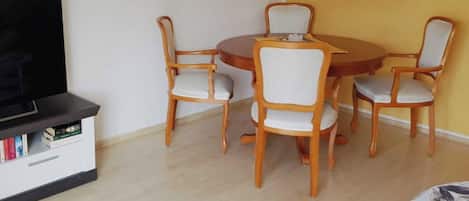 Un comedor con una mesa redonda de madera y seis sillas, un televisor sobre un mueble bajo y una estantería con libros.