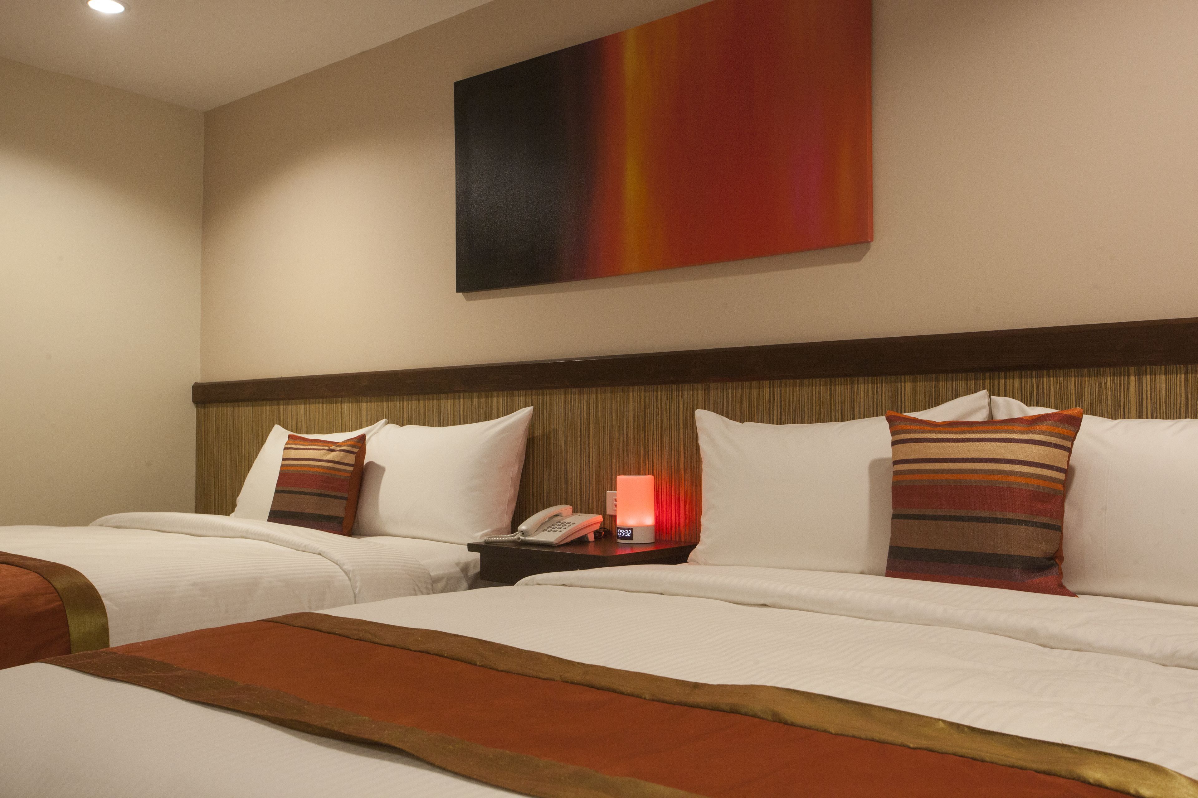 Une chambre d’hôtel avec deux lits, une table de chevet avec un téléphone et une lampe rouge, et un grand tableau abstrait accroché au mur.