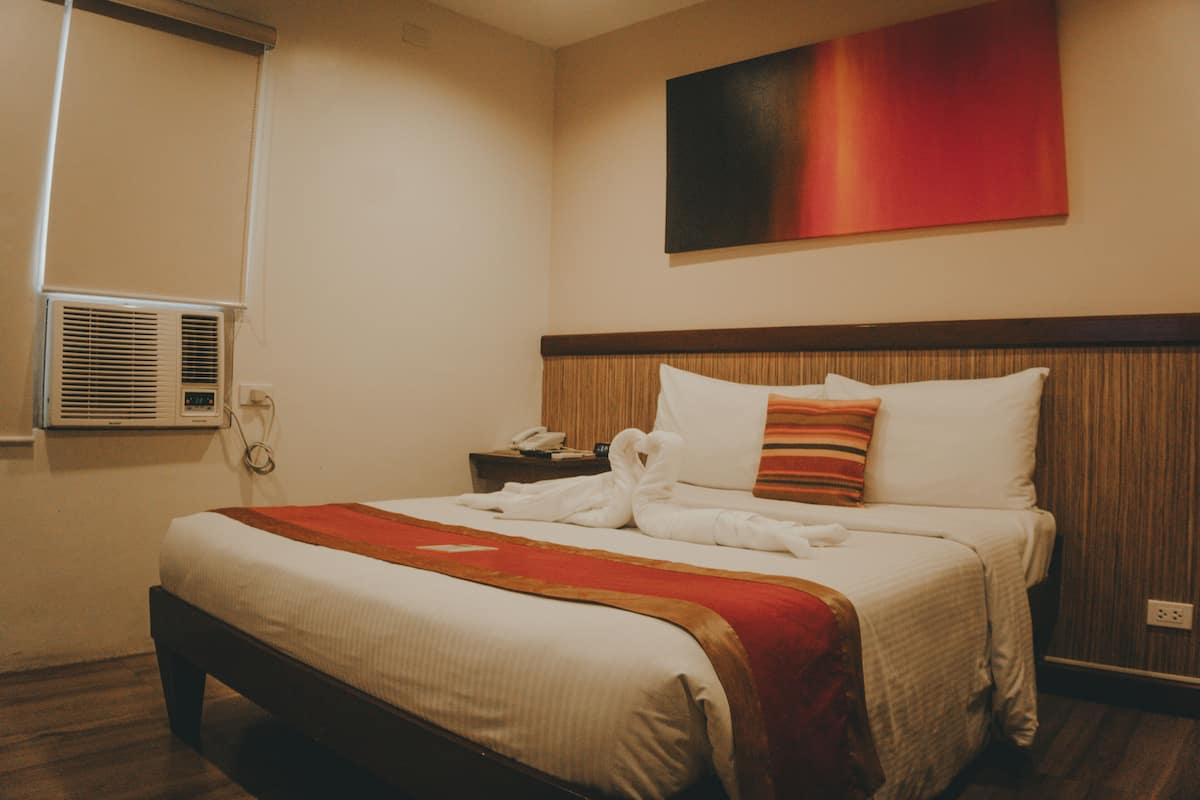 A hotel room with a large bed, a bedside table with a phone, a wall-mounted air conditioner, and a red abstract painting above the bed.