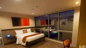 A hotel room with a large bed, a nightstand, a desk with a computer, and a window with a view of the city. - NS Royal Hotel (Cebu City)