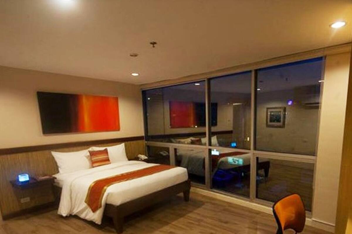 A hotel room with a large bed, a nightstand, a desk with a computer, and a window with a view of the city.