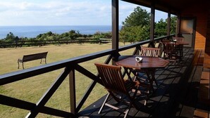 Un espace extérieur aménagé, abrité et doté de tables et de chaises en bois, offrant une vue sur un champ verdoyant et l’océan.