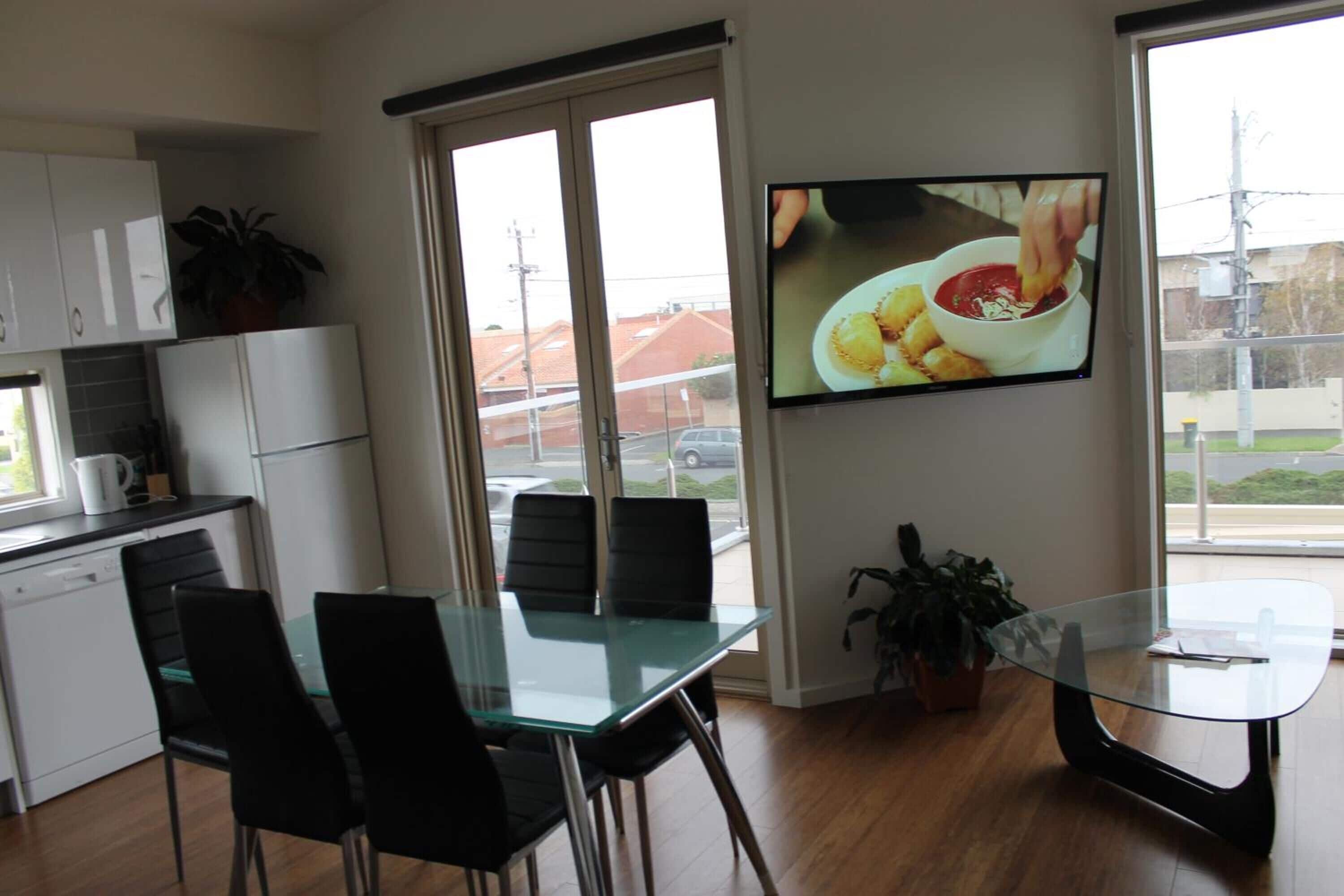 A modern kitchen with a dining area, a TV mounted on the wall, and a view of the outside.