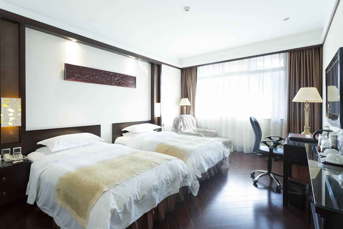 Ein Hotelzimmer mit zwei Betten, einem Schreibtisch, einem Stuhl und einem großen Fenster mit Vorhängen.