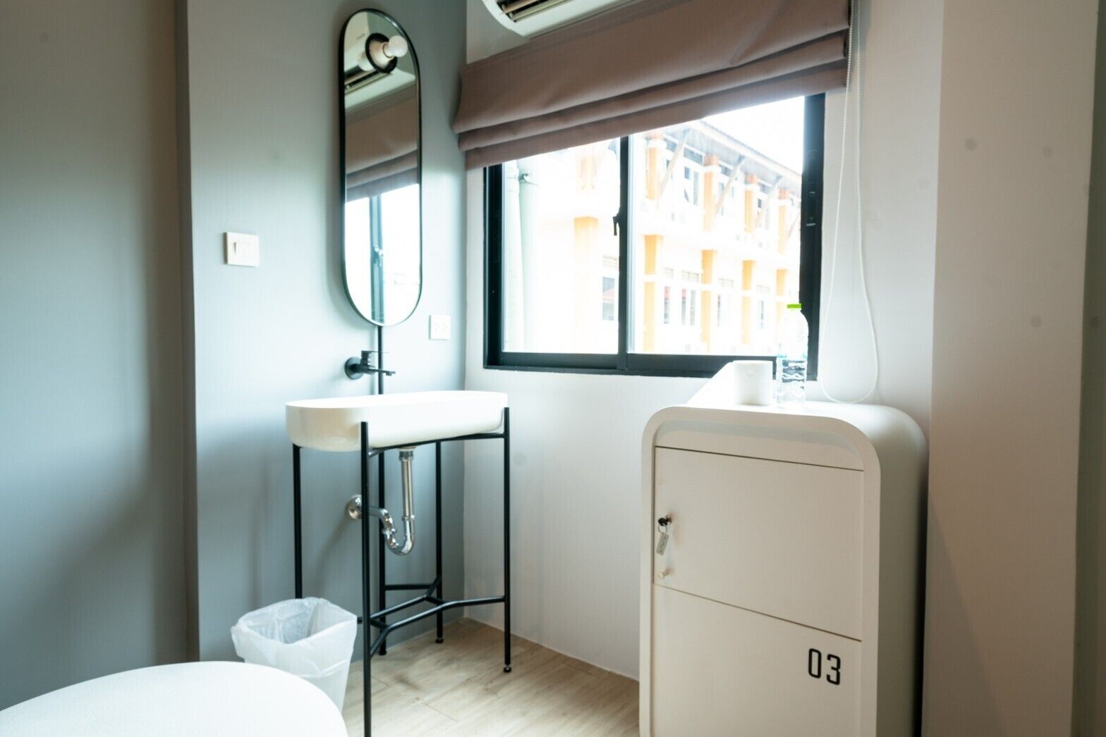 A small, modern room with a single bed, a white sink, a mirror, a window with a view of buildings, and a cabinet with the number 03.