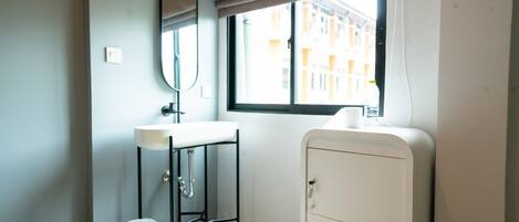 A small, modern room with a single bed, a white sink, a mirror, a window with a view of buildings, and a cabinet with the number 03.