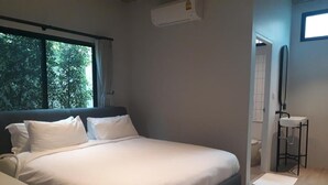 A bedroom with a bed, a window, an air conditioning unit, a bedside table, and a small bathroom with a toilet and sink. - Bedspread Hostel - Adults Only (Bangkok)