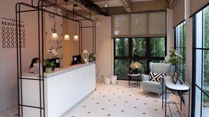 A modern reception area with a white desk, pendant lights, and a large window offering a view of greenery. - Bedspread Hostel - Adults Only (Bangkok)