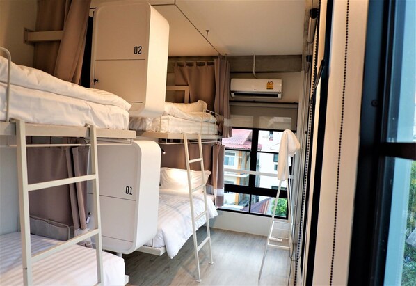 A dormitory-style room with bunk beds, numbered beds, and a window with a view of buildings outside. - Bedspread Hostel - Adults Only (Bangkok)