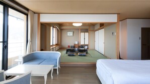 A hotel room with a sofa, a small table, a bed, and a futon bed. There is a window with curtains, a ceiling light, and a picture on the wall. - Kaisen no Yado Matsuura (Saiki)